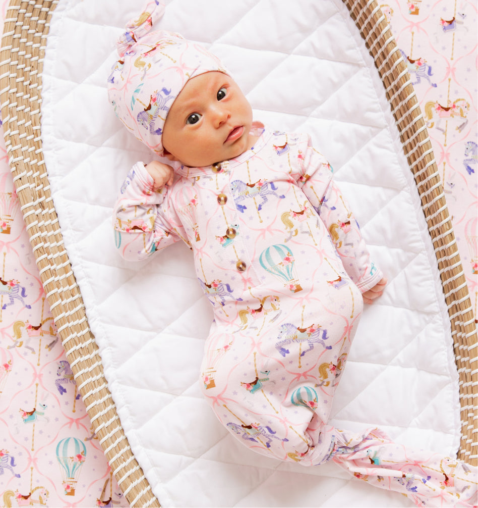 Newborn in pink knotted gown with carousel horse and hot-air balloon print, lying in wicker bassinet on quilted mattress.