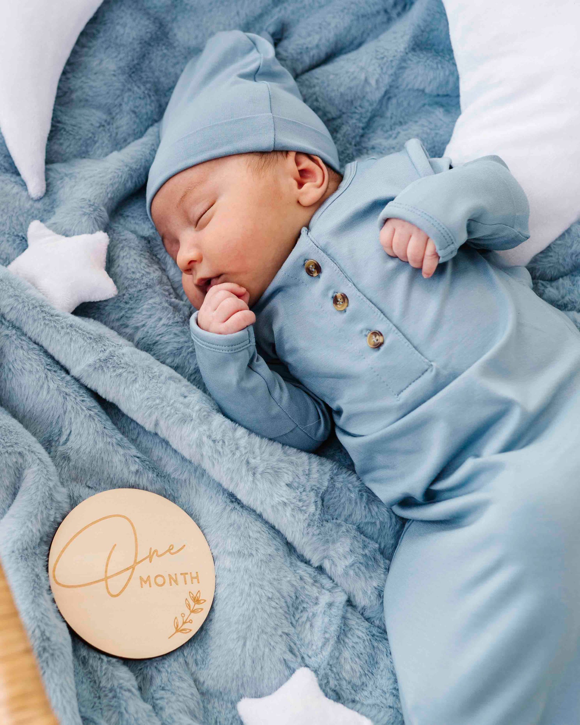 Sleeping newborn in blue pajamas on plush blanket with wooden milestone disk 