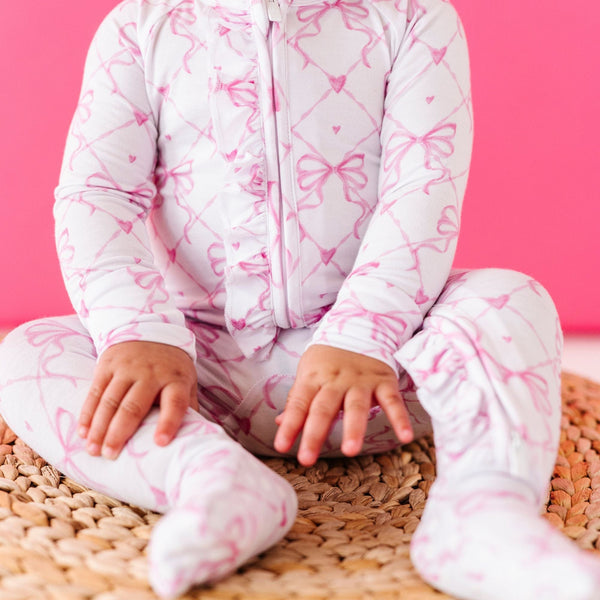 Baby in pink ribbon-patterned footed pajamas sitting on a woven mat, hands resting on knees