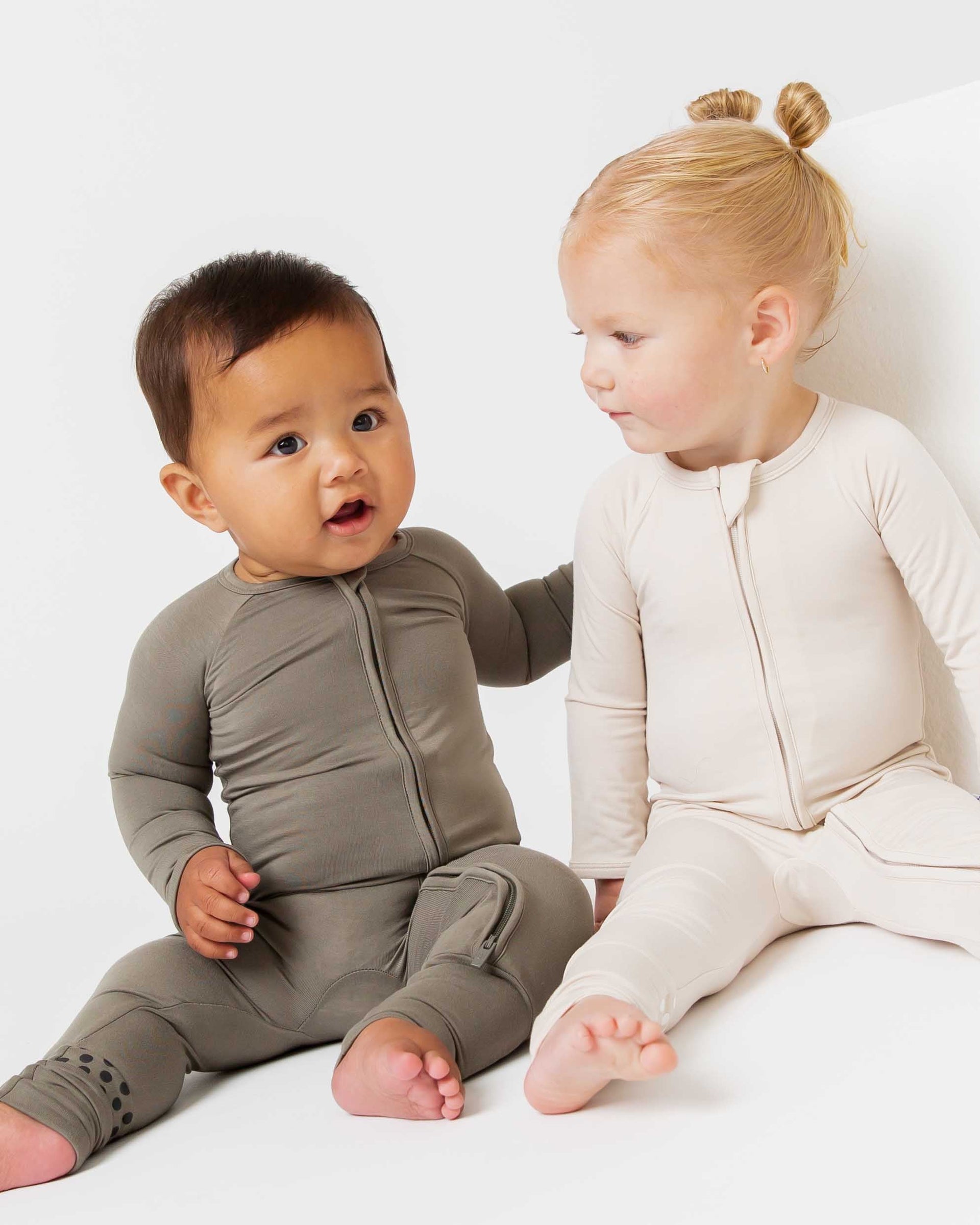 Two toddlers sitting wearing zip-front footless sleepers — one olive green, one cream