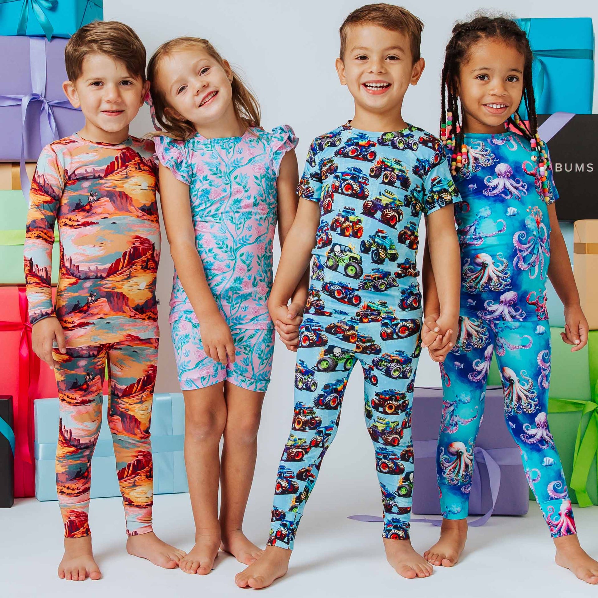 Four kids holding hands in colorful printed pajamas: desert, pink floral, blue trucks, blue octopus