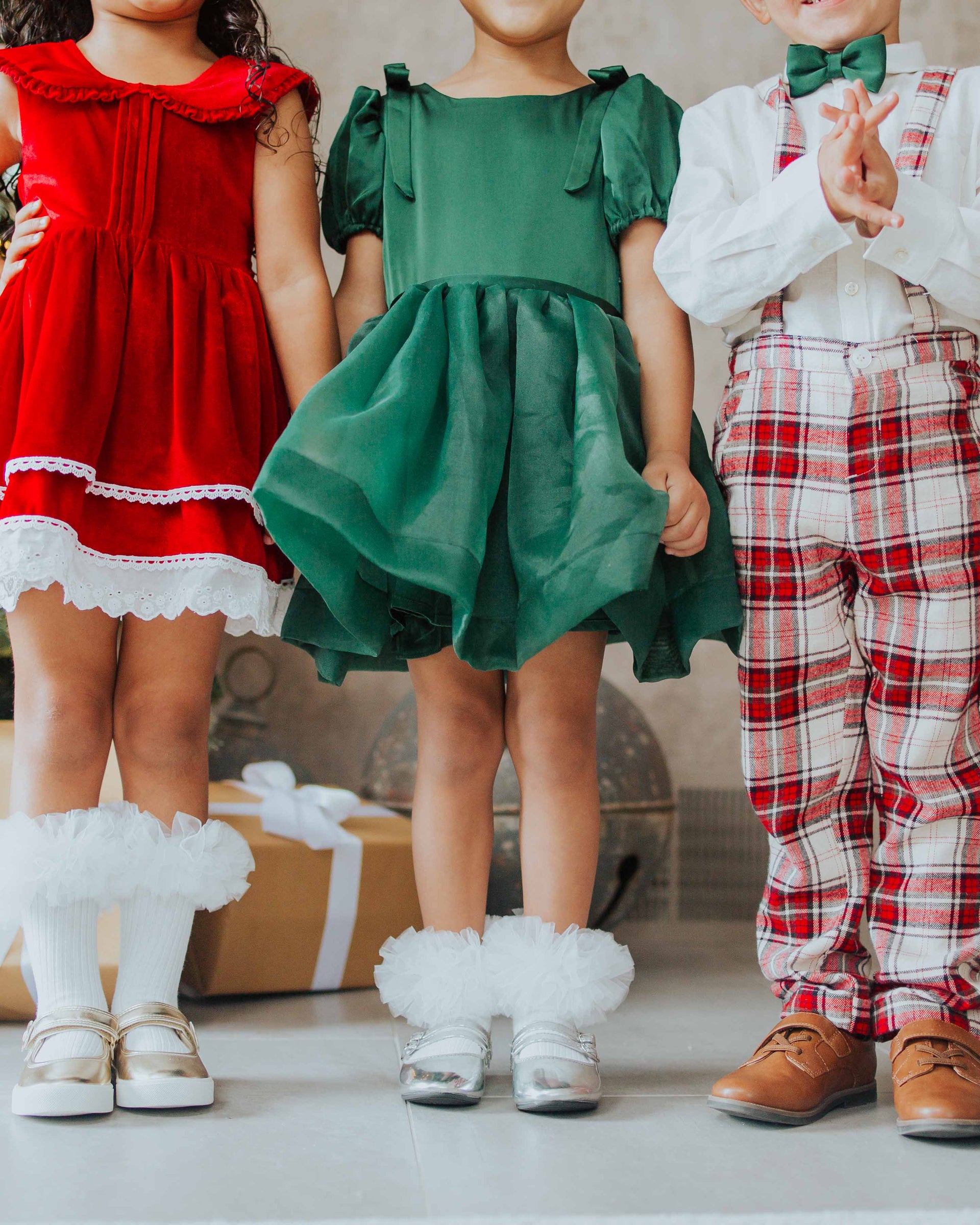 Three toddlers in holiday clothes: red velvet dress with lace, green puff-sleeve dress, and white shirt with red plaid pants.