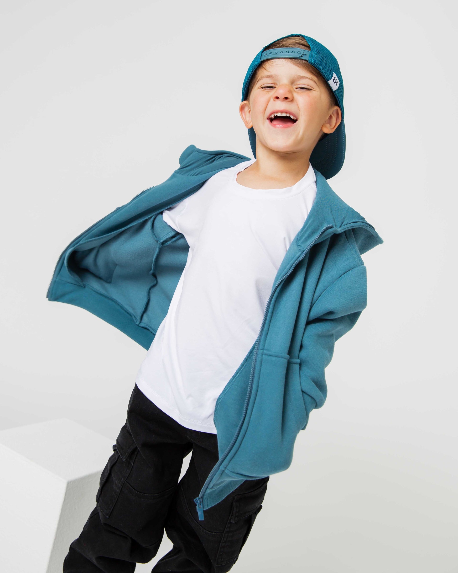 Smiling young boy wearing a teal zip hoodie, backward teal cap, white tee and black cargo pants.