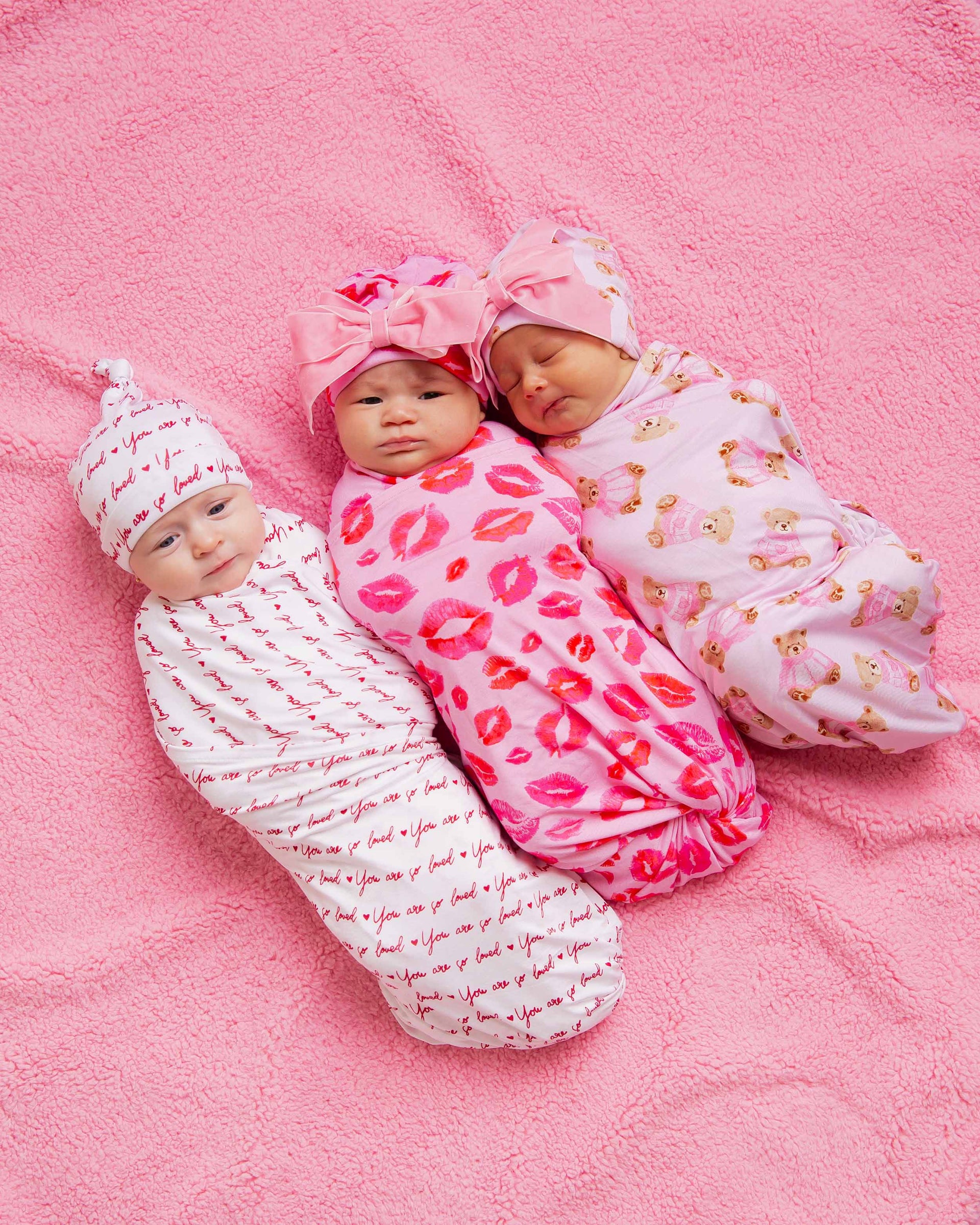 Three newborns swaddled on a pink blanket in bamboo wraps patterned with 'You are so loved', lips, and teddy bears