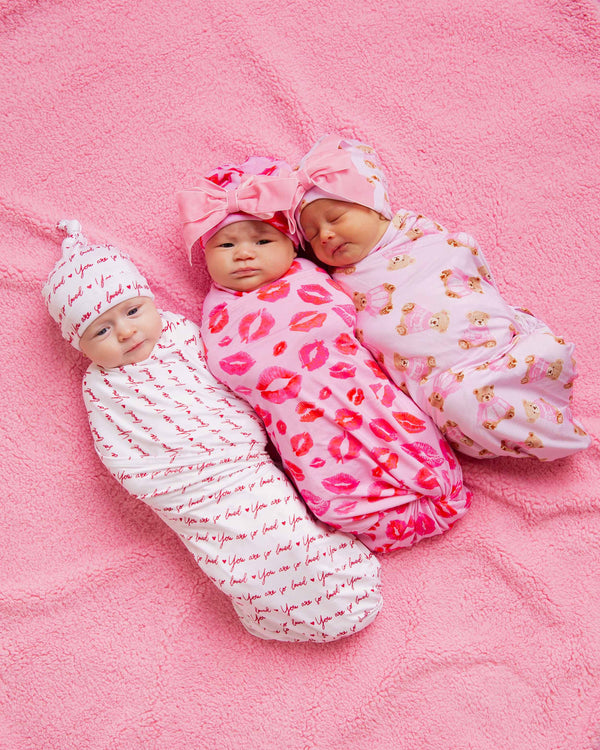 Three newborns swaddled on a pink blanket in bamboo wraps patterned with 'You are so loved', lips, and teddy bears