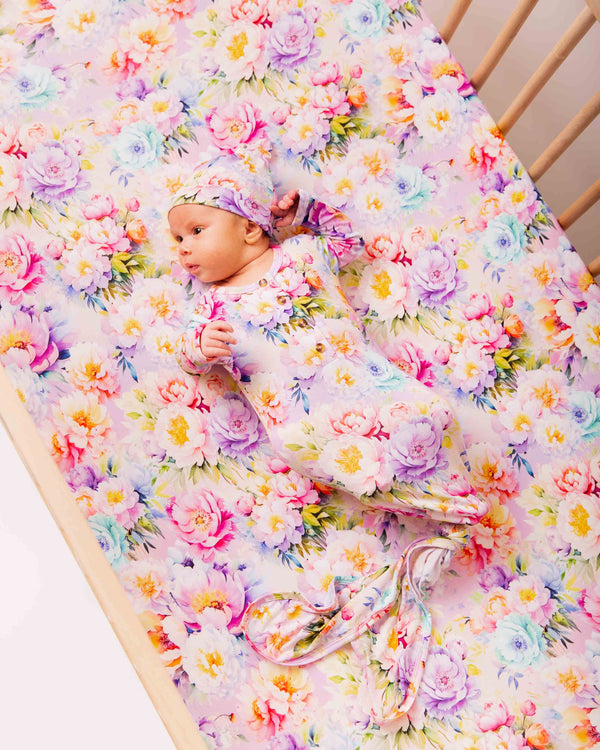 Infant lying in a wooden crib on pastel floral bamboo crib sheet, wearing matching hat and gown