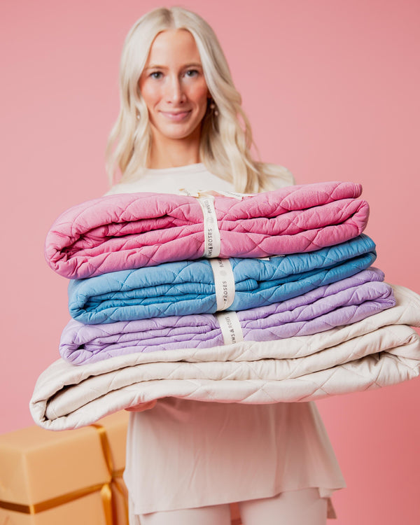 Woman holding folded cooling blankets in pink, blue, lavender, and cream against a pink background