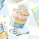 A colorful dessert in a glass cup with rainbow layers and whipped cream on top.